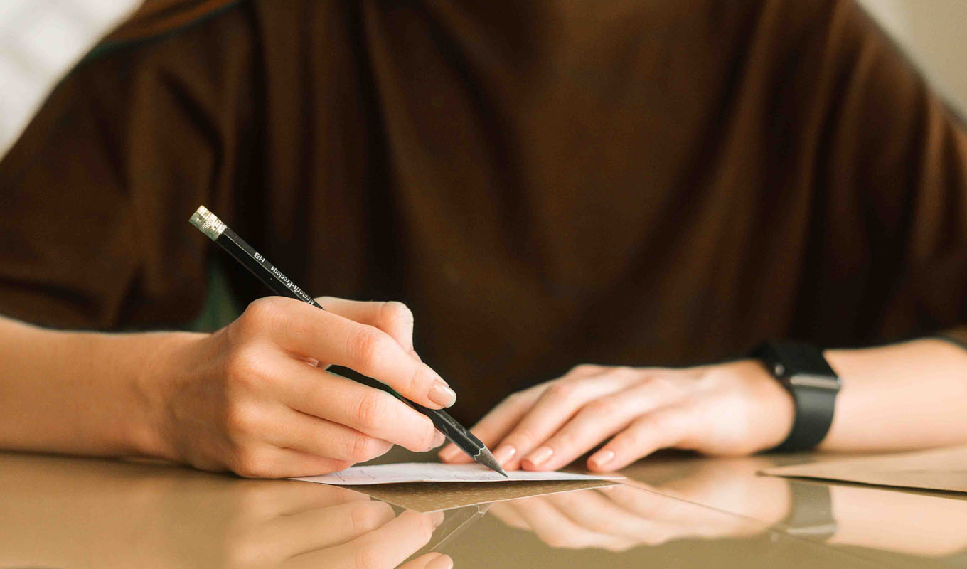 Close-up of a person’s hands writing a handwritten note with a pencil on paper at a table.