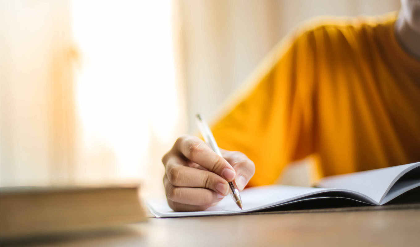 A person wearing a short‑sleeved shirt writes in an open notebook with a pen at a table, with a closed book placed nearby and soft, warm light coming from the background.