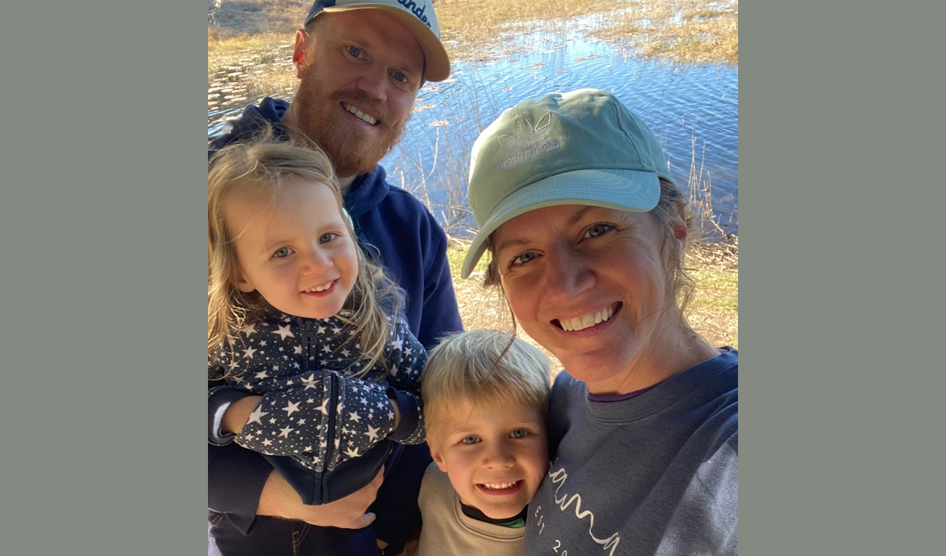 A smiling family of four—two adults and two young children—taking a selfie outdoors in front of a pond with tall grasses on a sunny day.