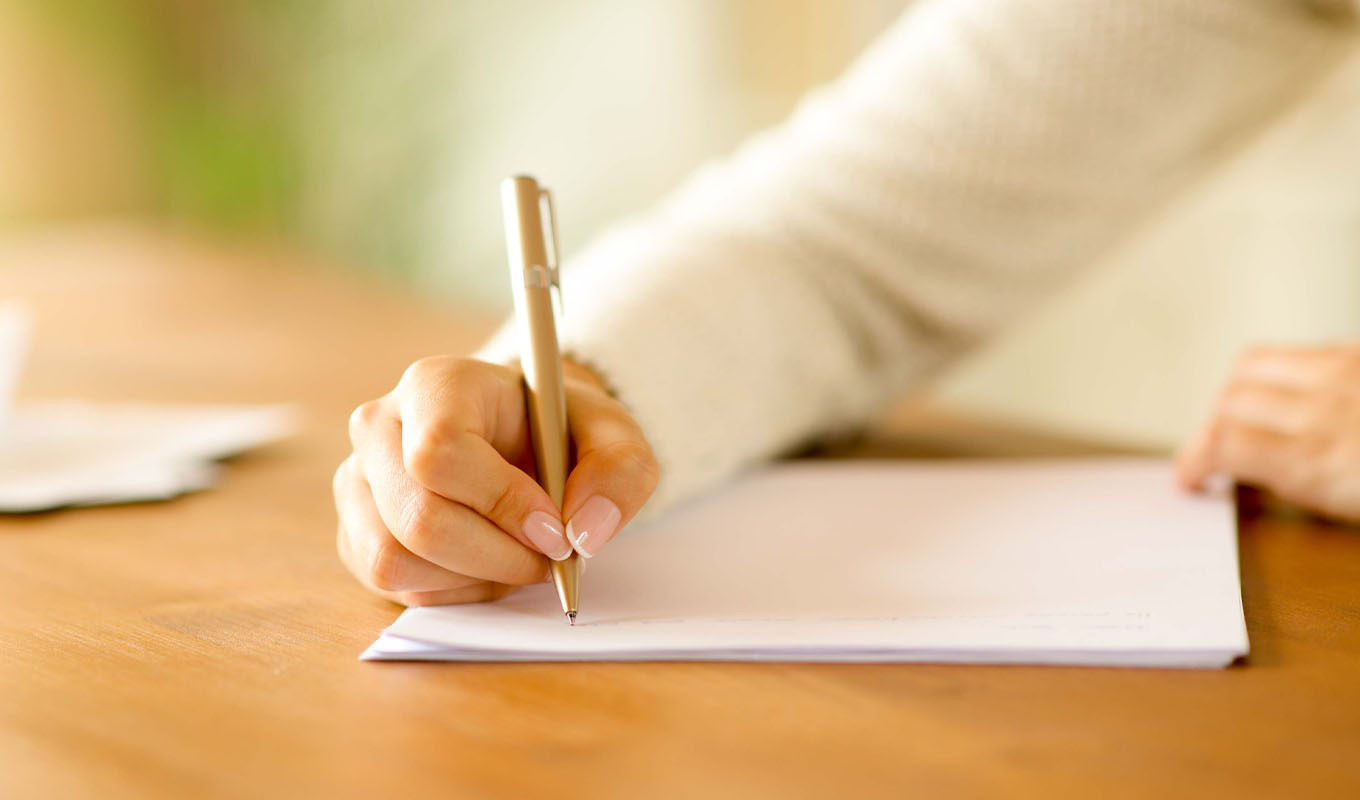 Close‑up of a person writing a letter with a pen on paper at a wooden desk, representing member communication, personal correspondence, or written outreach.