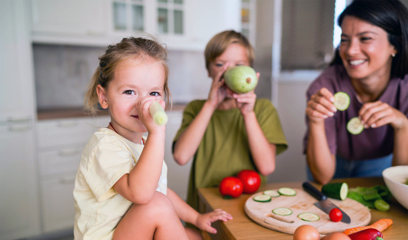An adult and two children sit at a kitchen table preparing fresh vegetables, including cucumbers, tomatoes, and carrots, with a cutting board and kitchen knife visible in a bright home kitchen.