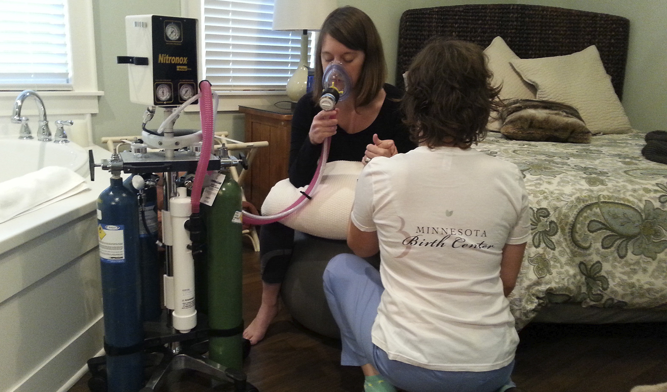 A woman in labor sitting on a bed inhales nitrous oxide while another woman kneels down to assist her