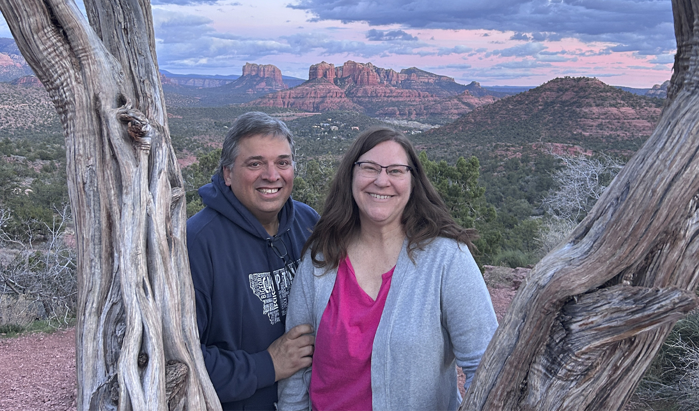 A man and woman stand together, smiling in front of a large, leafy bush and a desert backdrop, enjoying a sunny day outdoors.