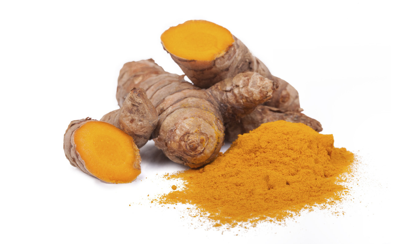 Several turmeric roots, one cut to reveal its bright orange interior, are arranged beside a pile of ground turmeric powder on a white surface.