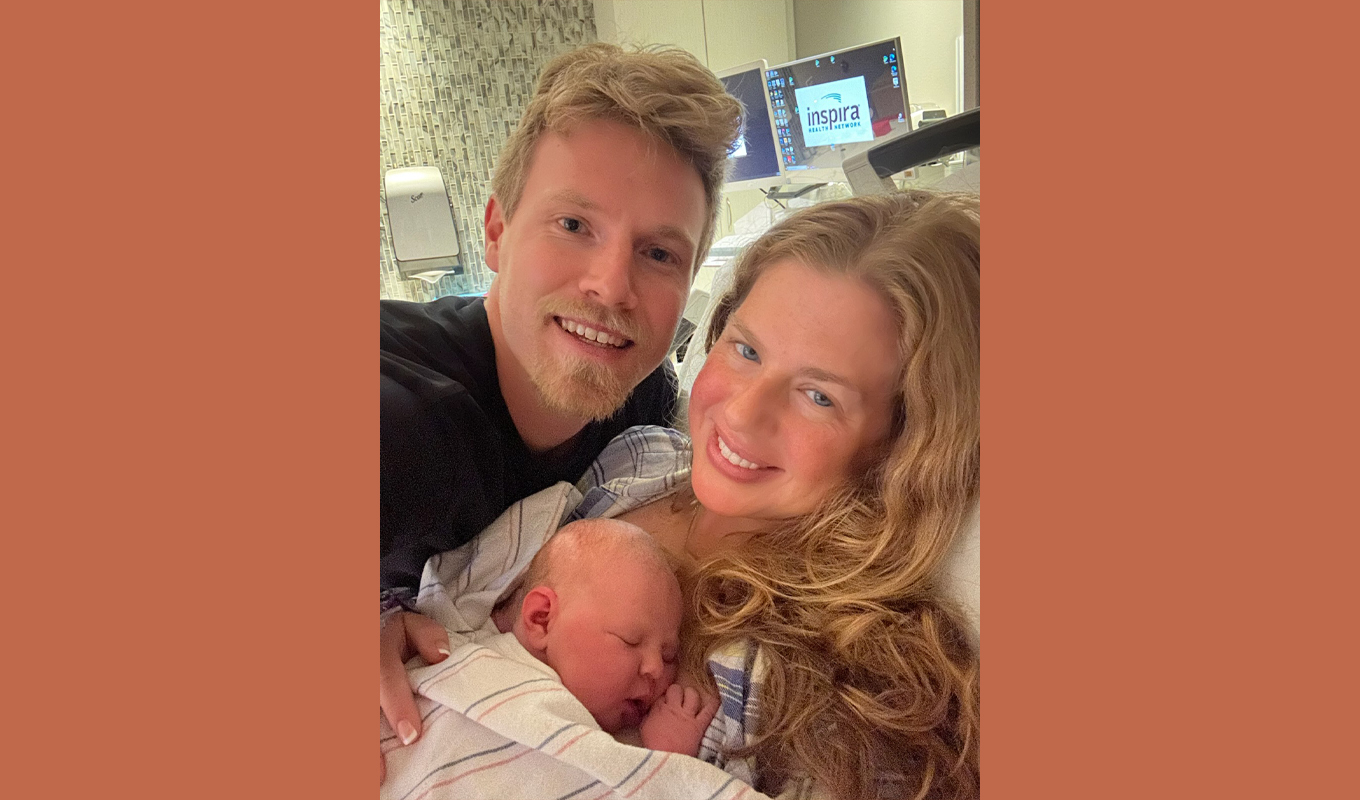 Newborn wrapped in a striped blanket held by two adults in a hospital room with medical equipment visible.