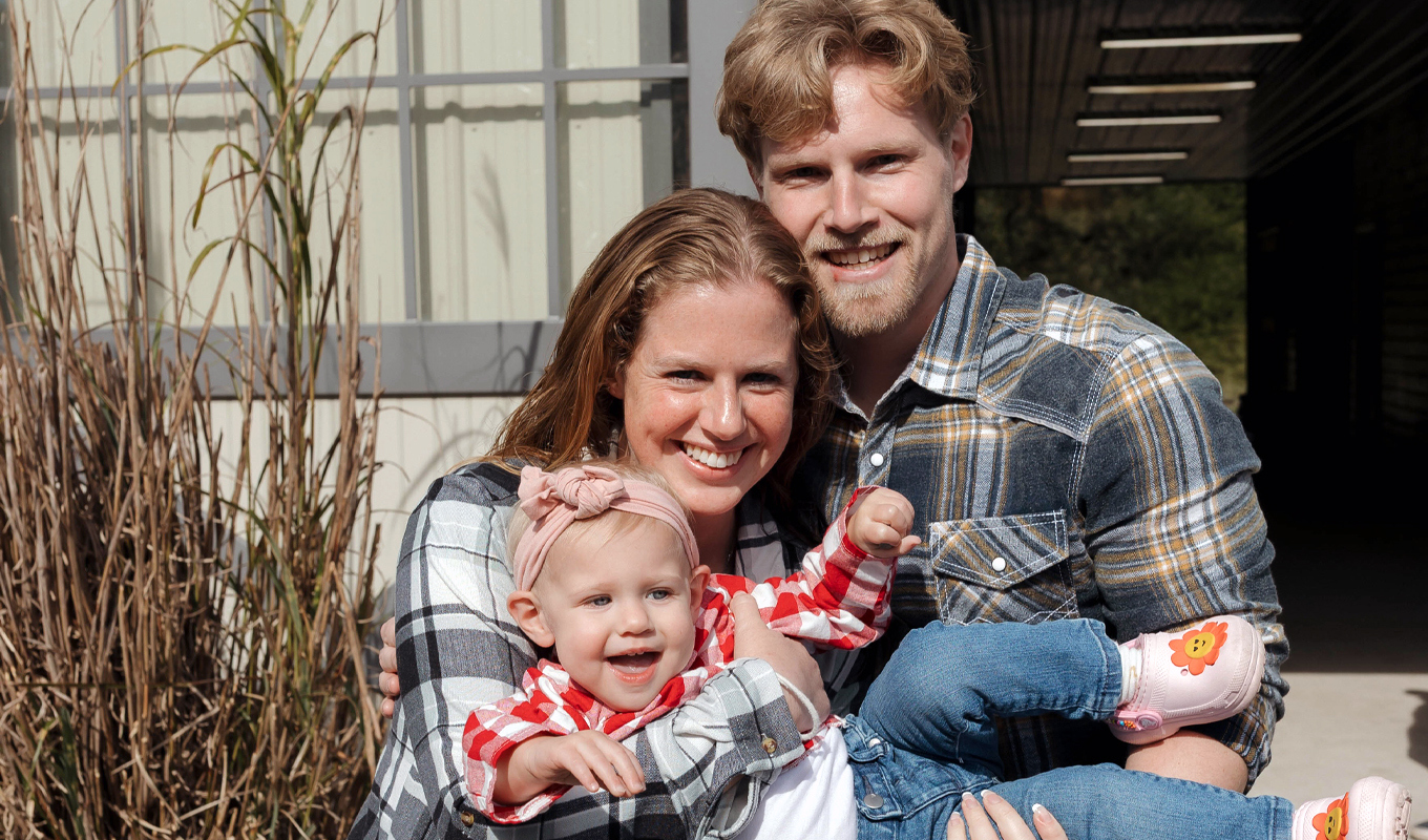 Two adults are holding a small child dressed in a red-and-white checkered shirt, blue jeans, and pink shoes with flower designs.