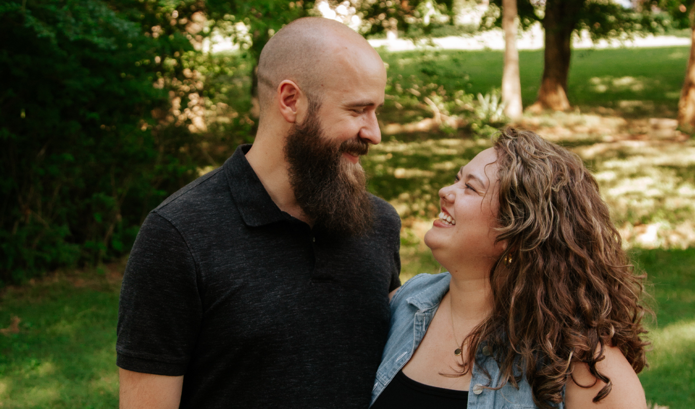 Two people standing close together outdoors in a grassy, tree‑filled area, with one person turned slightly toward the other in a relaxed, natural pose.