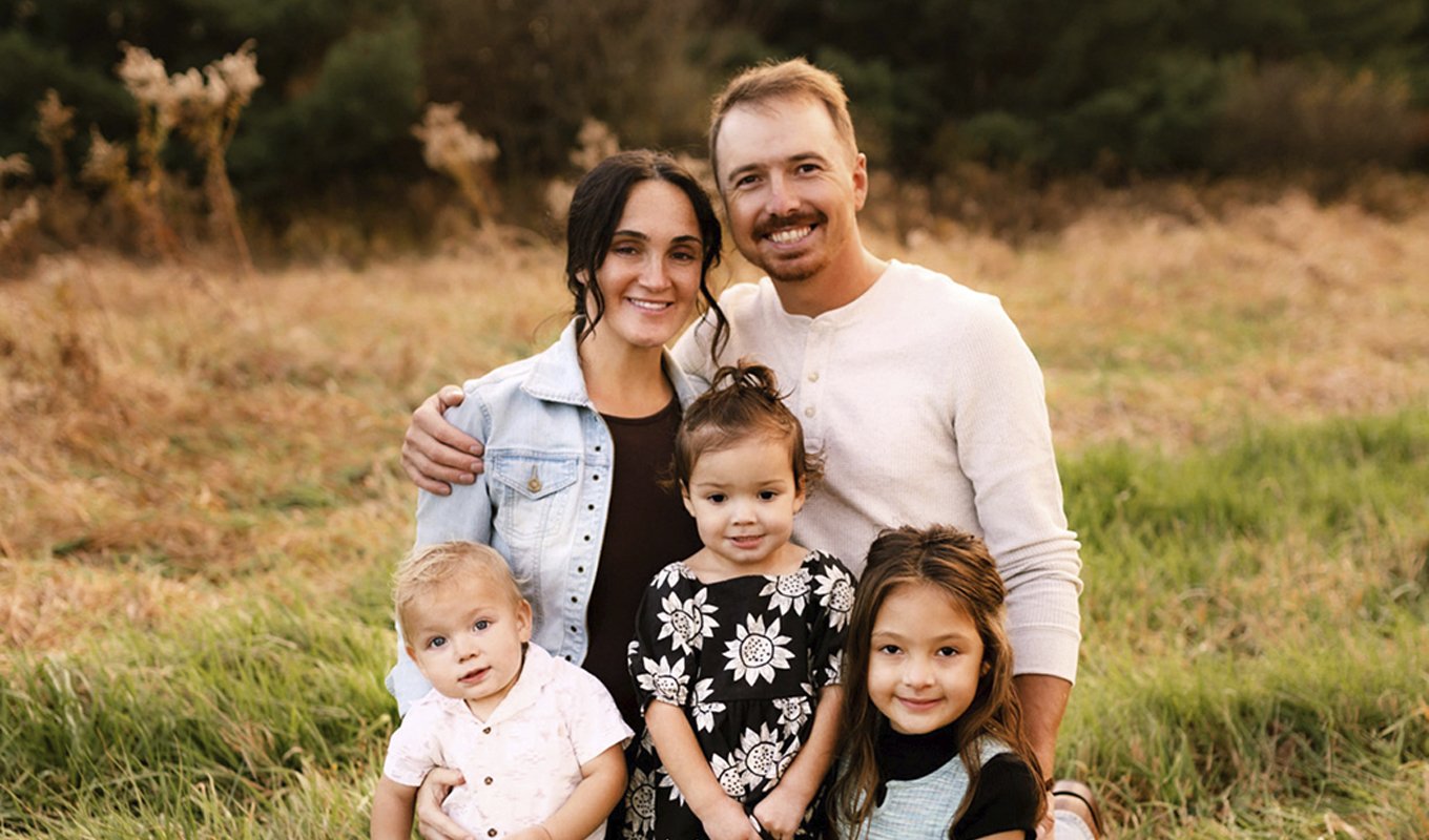 A happy family of five posing outdoors in a grassy field during sunset. Two adults and three young children smile at the camera, dressed in casual clothing.
