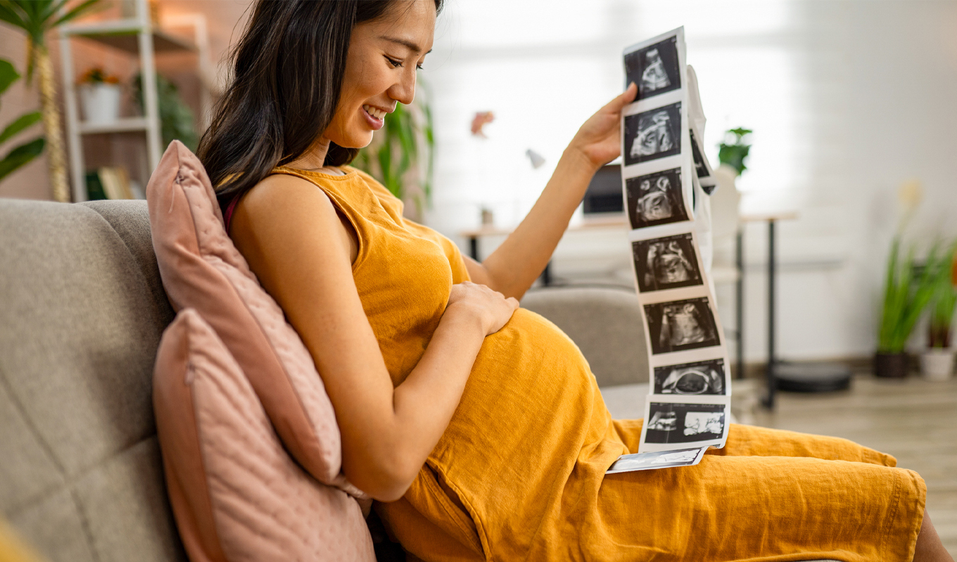 A person sitting on a couch, wearing a loose sleeveless dress and holding a long strip of ultrasound images while resting a hand on their abdomen in a bright, comfortable living room.