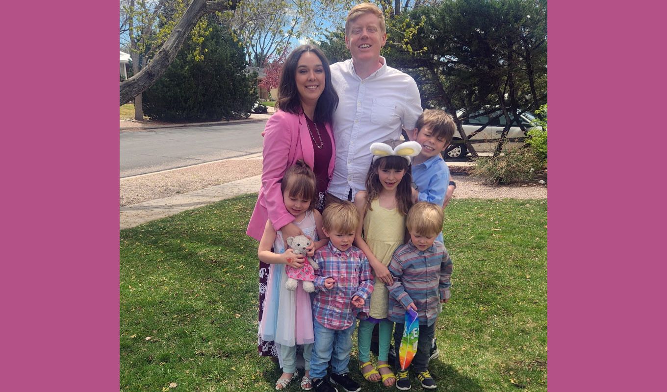 A mother, father, and children stand together outdoors on a grassy lawn, wearing spring clothing, with trees and a residential street in the background.