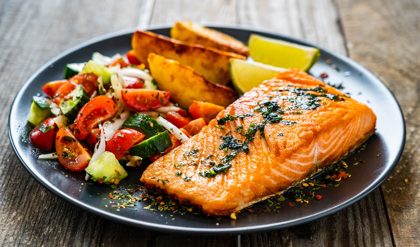 A plate with a seared salmon fillet garnished with herbs, served alongside roasted potato wedges, a fresh tomato‑cucumber salad, and lime wedges on a wooden table.