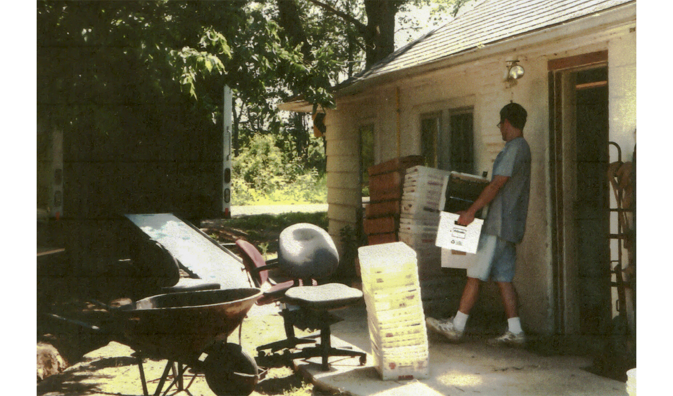 A man stands outside a house, holding a box, with a clear sky in the background and greenery surrounding the property.