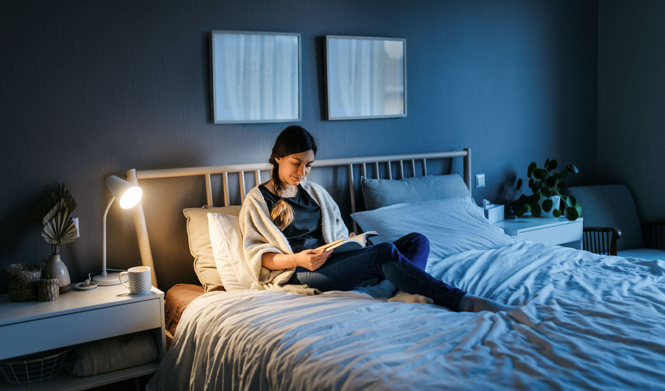 Person sitting on a bed in a softly lit bedroom, reading a book next to a bedside lamp and nightstand.