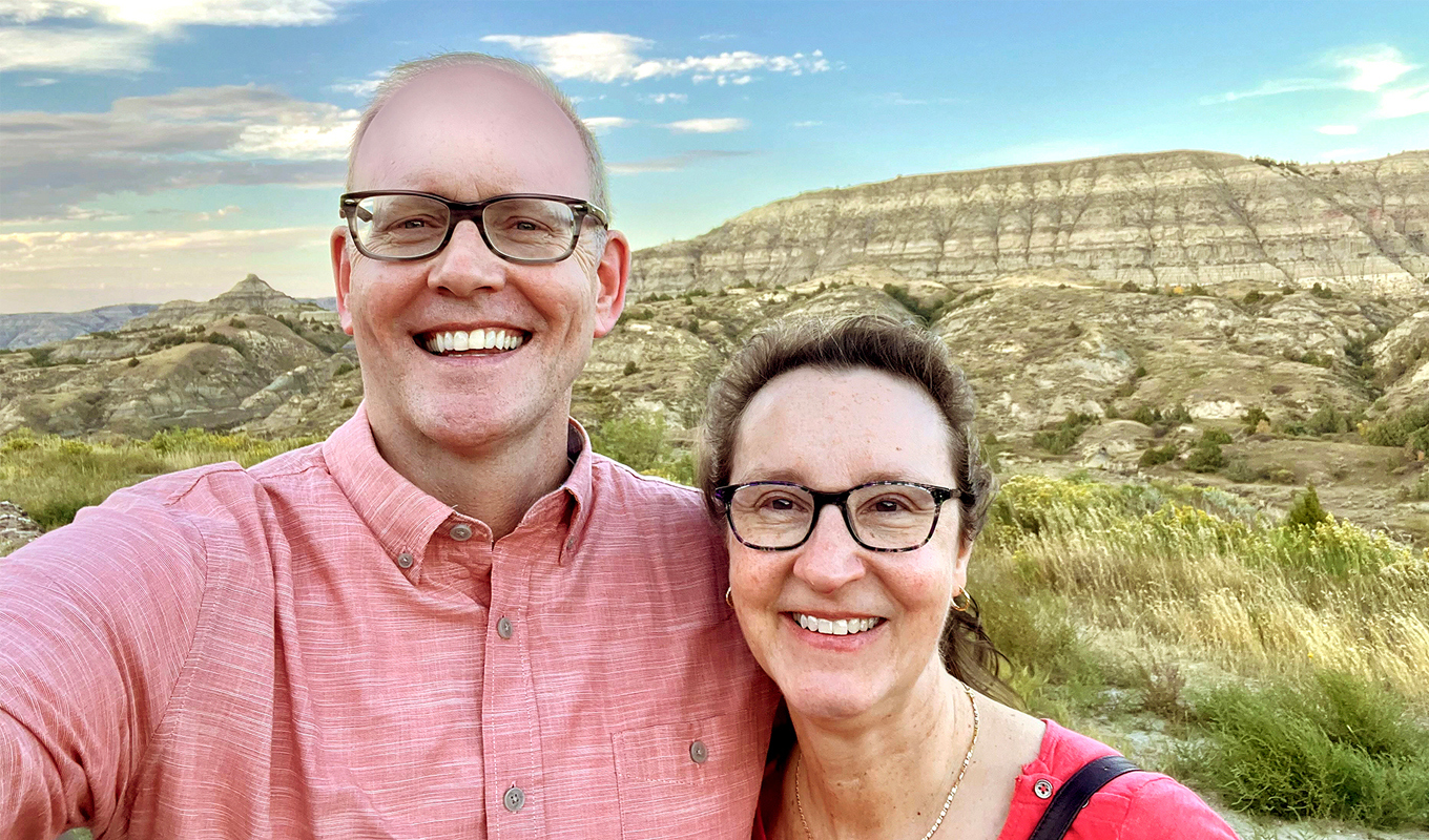 Two smiling individuals taking a selfie with the scenic Badlands National Park in the background.