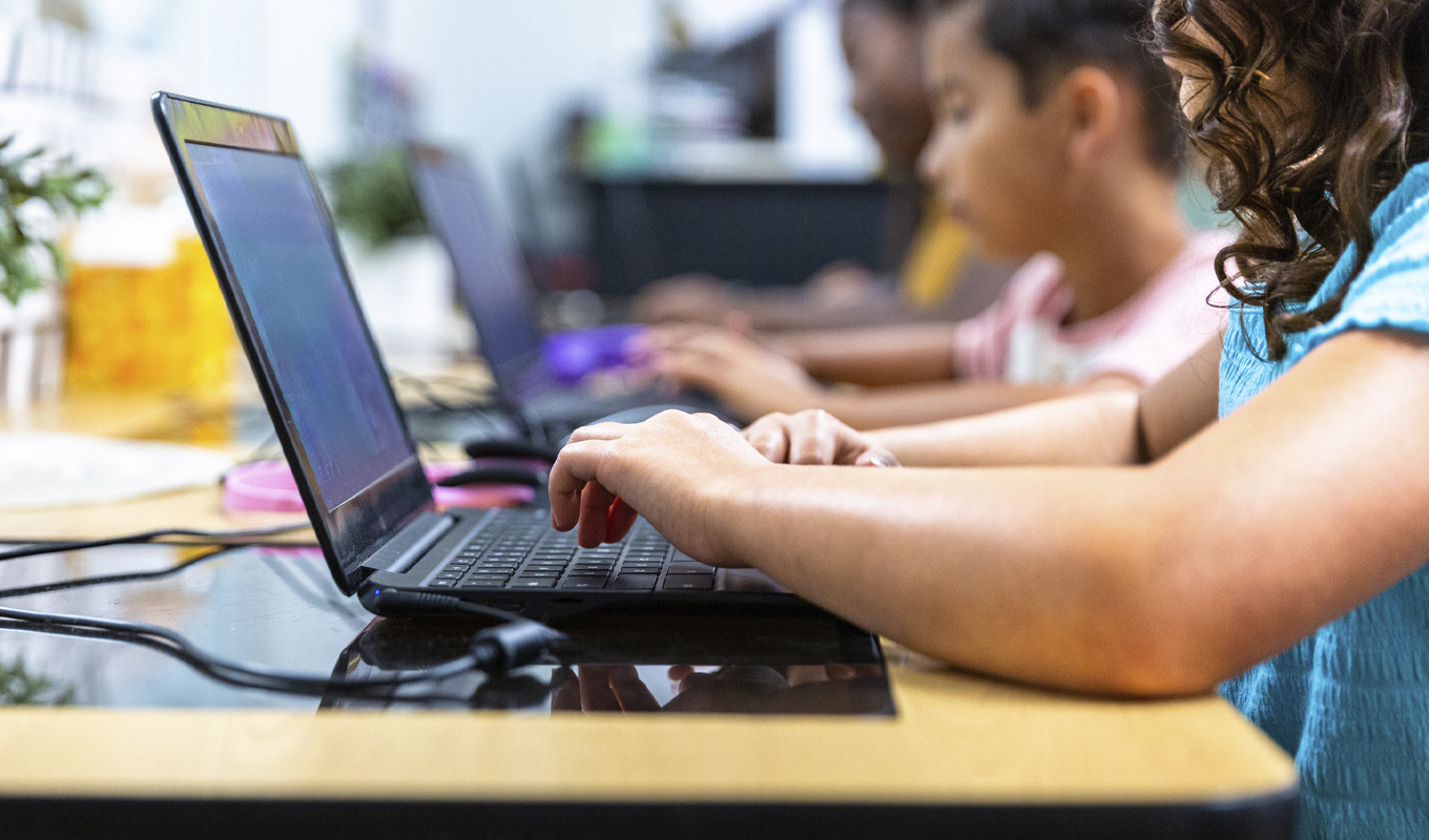 Grade-school students sit at their desks typing on laptop coputers.