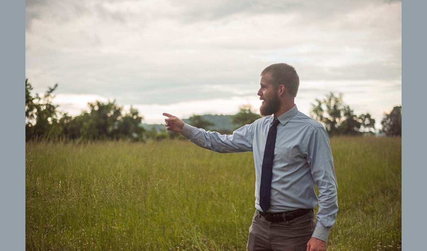 Man wearing a dress shirt and tie standing in tall grass, pointing across a field.