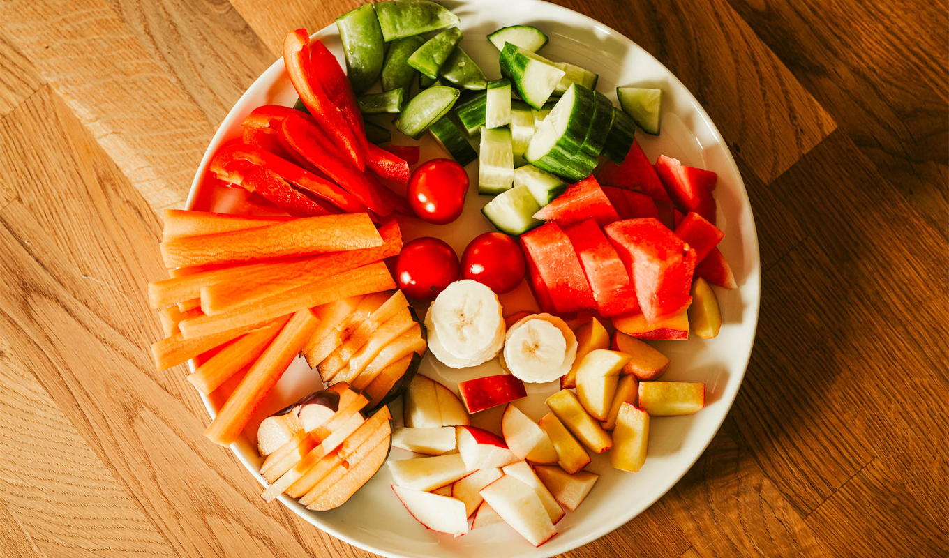 A round plate on a wooden table arranged with sliced fresh produce, including carrots, red bell peppers, cucumbers, cherry tomatoes, watermelon, apples, and banana pieces, organized in colorful sections.