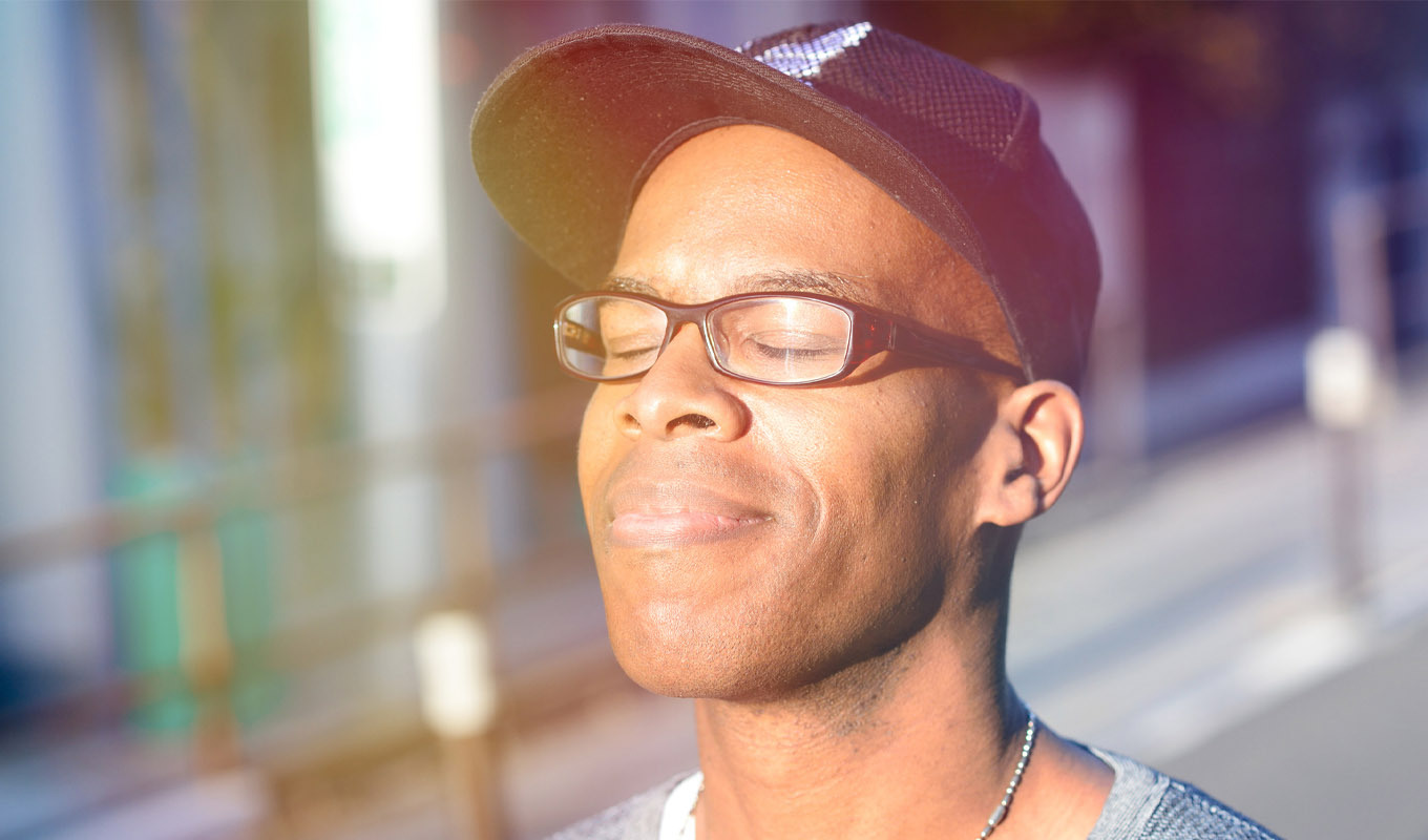 A person wearing glasses and a dark cap stands outdoors with eyes closed, face turned toward bright sunlight, with a sunlit building and railings blurred in the background.