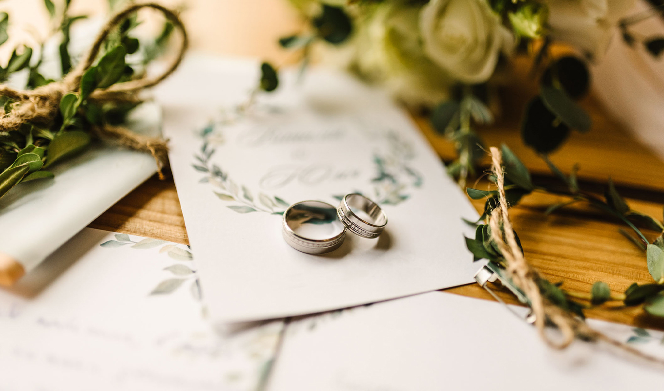 Two silver wedding rings placed on a floral wedding invitation, surrounded by greenery, twine, and soft decorative elements on a wooden table.