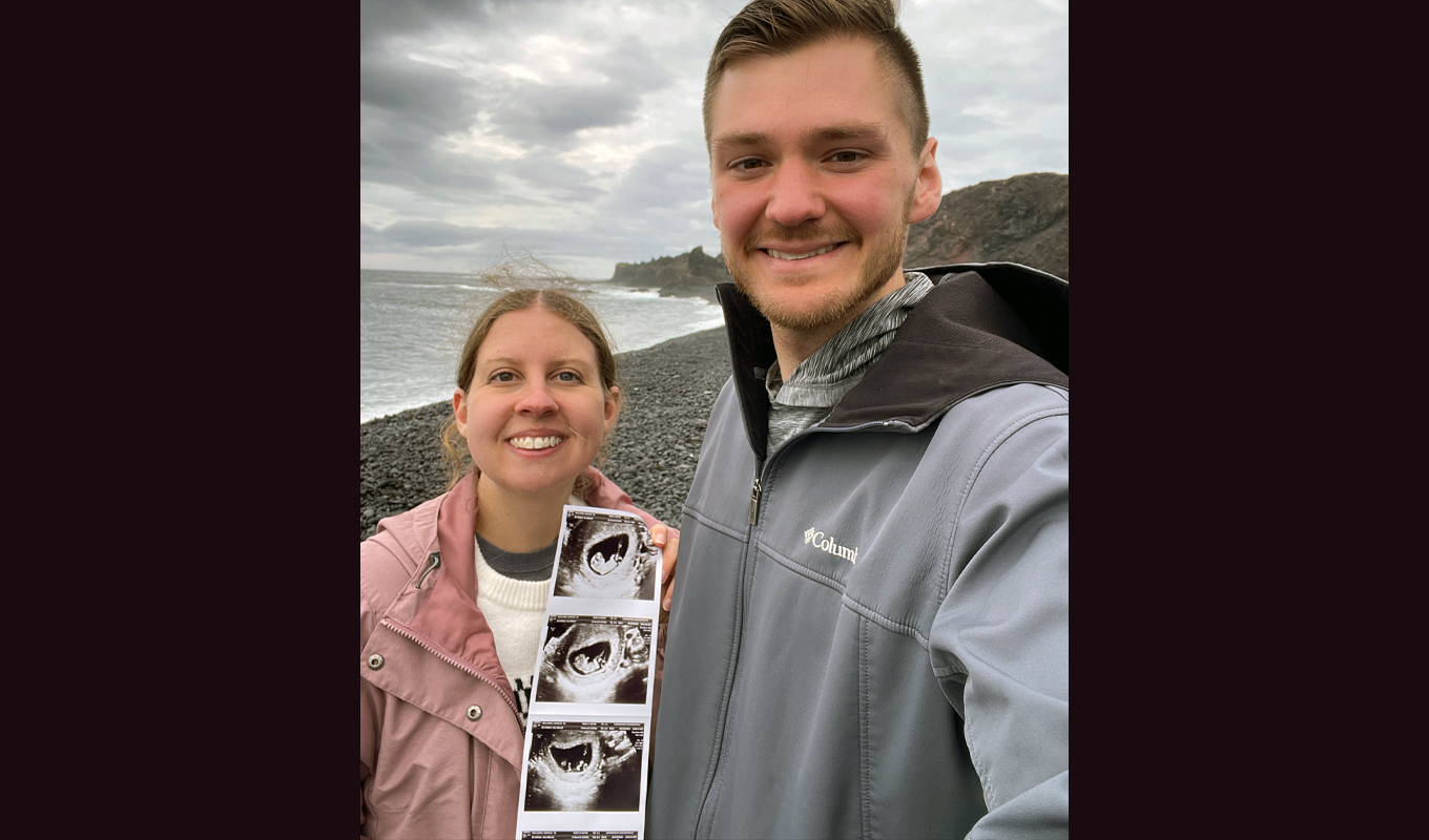 Two people standing on a rocky beach holding ultrasound images with ocean and cloudy sky in background.